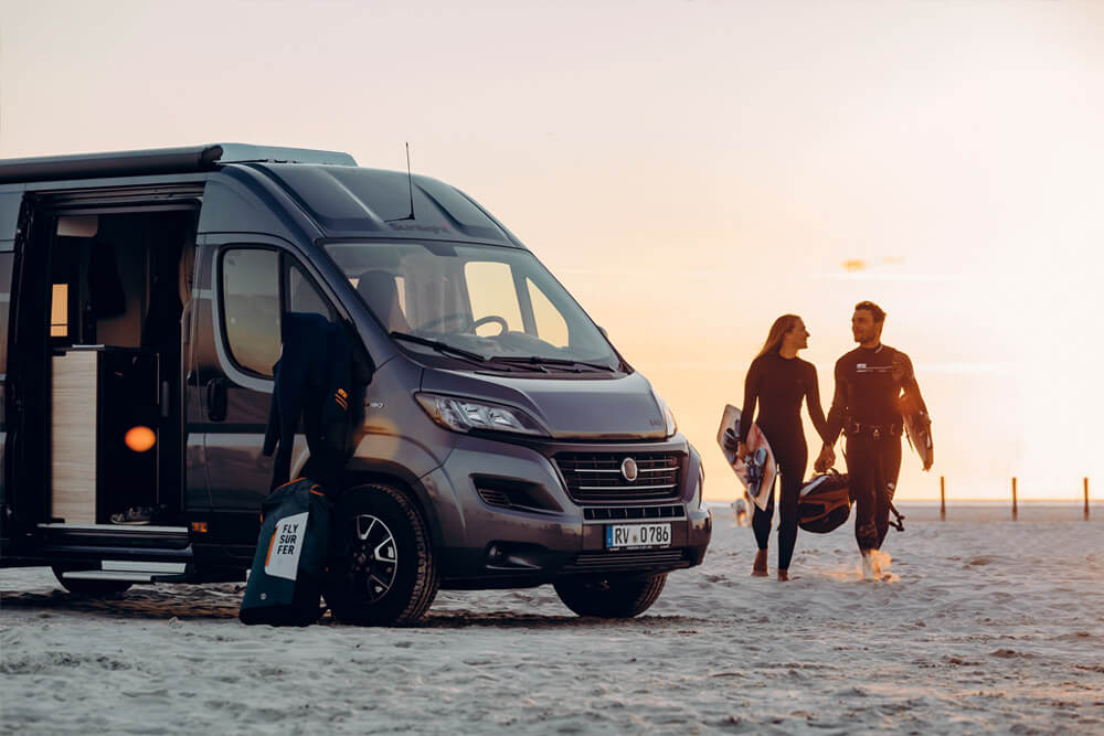 Wohnmobil am Strand bei einem Sonnenuntergang mit zwei Personen und Surfbretter