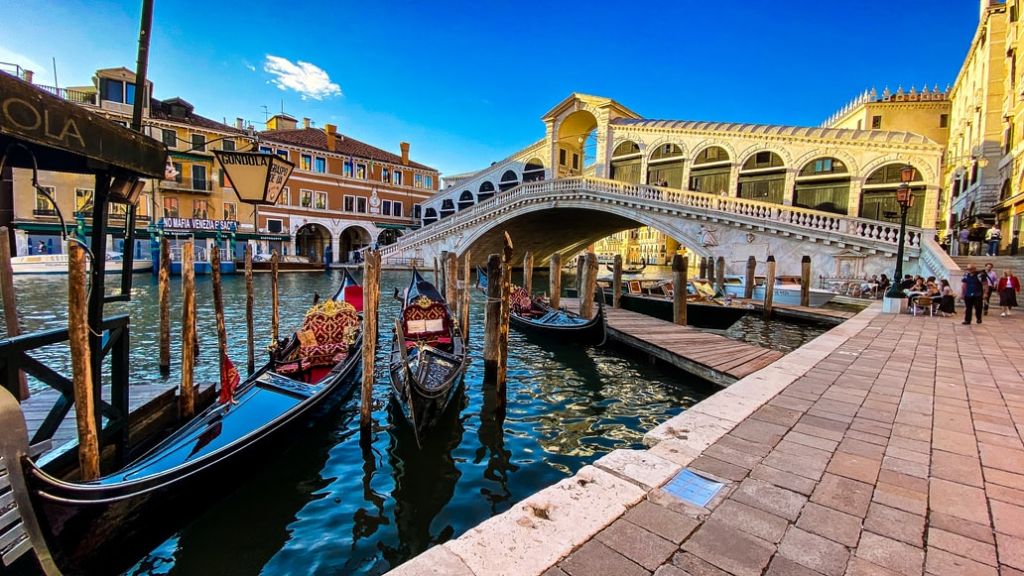 Während ihres Wildcamping Roadtrips durch Italien sollten Sie auf jeden Fall einen Abstecher in die Stadt der Gondeln und Liebe machen. Im Hintergrund ist die Rialtobrücke abgebildet. Im Vordergrund einige Gondeln in den schmalen Wasserkanälen von Venedig.