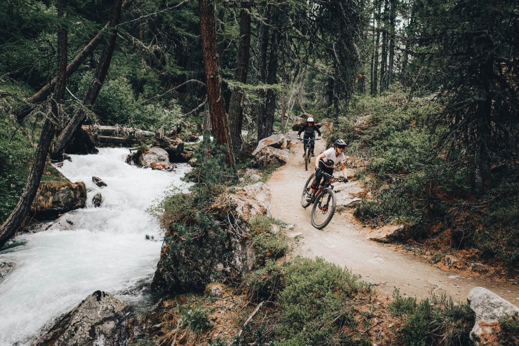 Bikepark Camping - zwei Mountainbiker fahren durch den Wald an einem Fluss vorbei