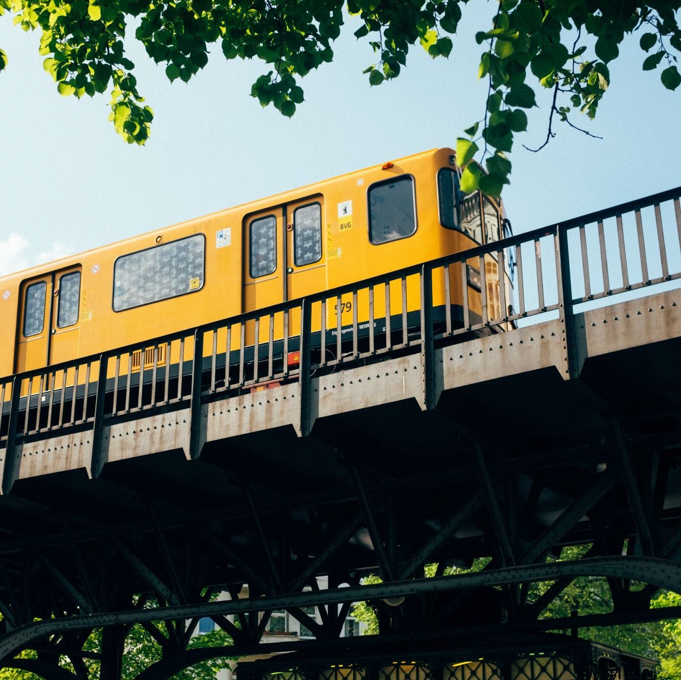 Gelbe Straßenbahn in Berlin fährt auf einer Brücke. Oben im Bild hängen Äste