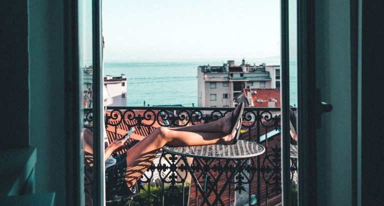 Überwintern in Südfrankreich - Frau sitzt auf einem Balkon mit Blick auf das Meer. Sie hat Ihre Füße auf einem kleinen Tisch hochgelegt und hält eine Zeitschrift in den Händen.