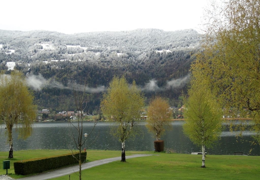 Camping Österreich mit Hund - Blick auf den Ossiacher See in Kärnten mit schneebedeckten Bergen und Bäume im Hintergrund.