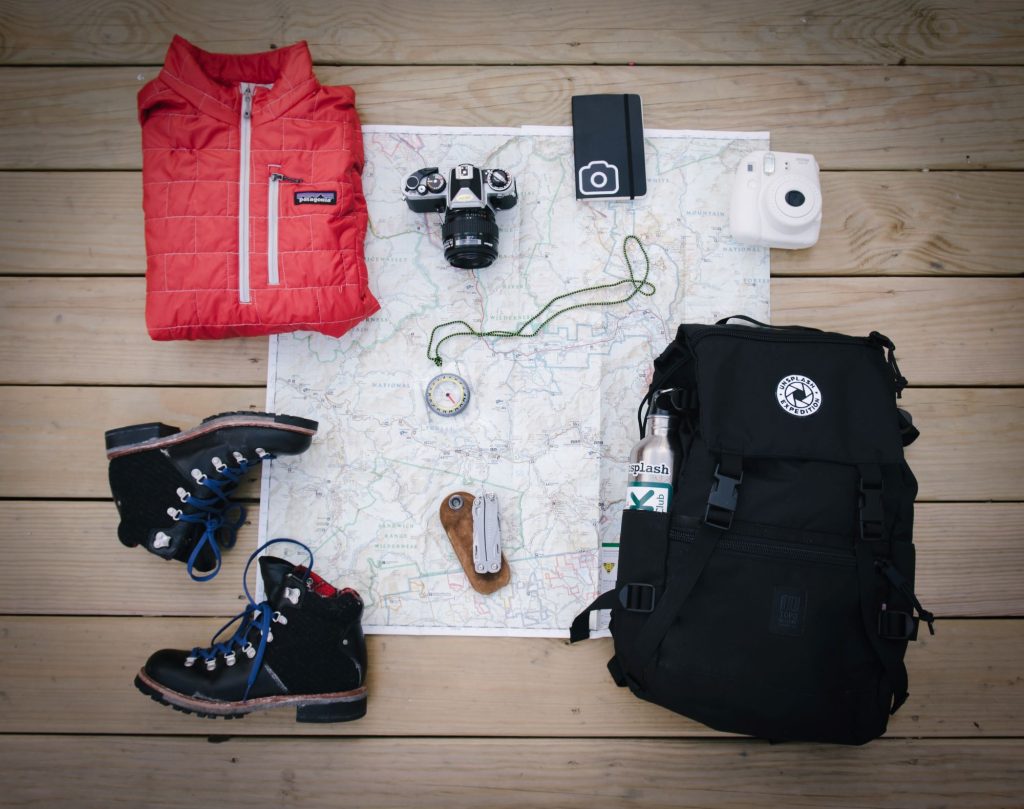 Nachhaltig Campen - Blick auf eine Karte, eine Kamera, Schuhe, einen Rucksack, ein Notizbuch, eine Jacke, einen Kompass und ein Taschenmesser.