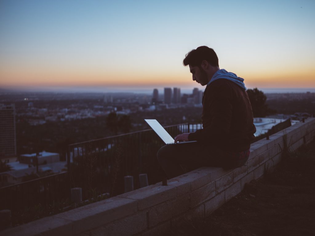Bester WLAN-Router für Wohnmobil - Mann sitzt auf der Kante eines Häuserdachs und guckt auf seinen Laptop. Im Hintergrund die Skyline einer Stadt. 