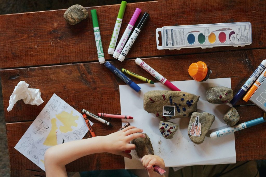 Bastelmaterial liegt auf einem Tisch. Ein Kind malt gerade einen Stein an