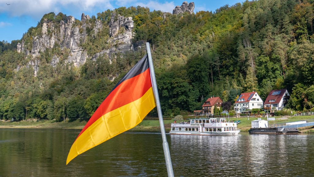 Camping Sächsische Schweiz - im Vordergrund die deutsche Flagge und dahinter ein Fluss und eine Steilklippe mit Bäumen.