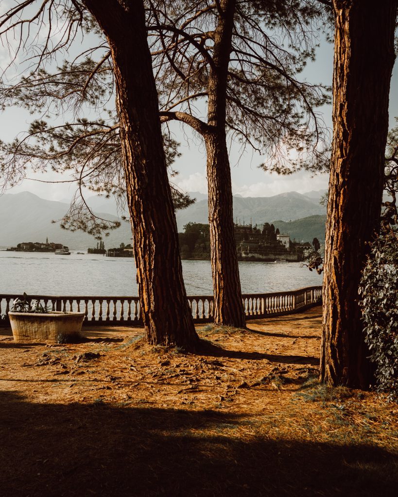 Camping Lago Maggiore mit Hund - Die Terrasse einen Hauses am See mit Pinienbäumen und einem altertümlichen Geländer. Dahinter der See und Wald.