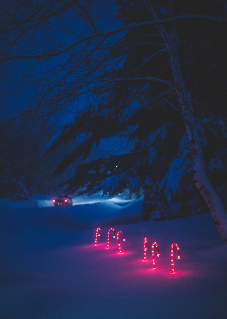 Weihnachten im Wohnmobil - Ein verschneiter Winterwald bei Nacht mit leuchtenden, roten Lichterketten, die die Form von Zuckerstangen haben und in den Schnee gesteckt sind, während im Hintergrund die Scheinwerfer eines Fahrzeugs sichtbar sind.
