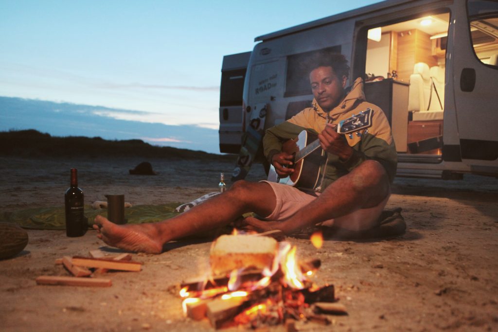 Wildcamping Strafen - Blick auf einen am Boden sitzenden und Gitarre spielenden Mann mit einem Wohnmobil im Hintergrund sowie einem kleinen Lagerfeuer im Vordergrund.